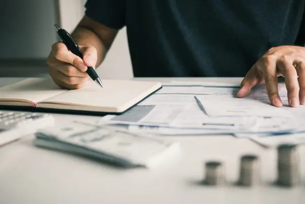 Person schreibt in Notizbuch, umgeben von Rechnungen, Taschenrechner und Münzen auf einem Tisch.