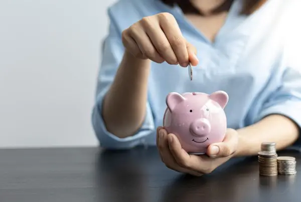 Hand legt Münze in rosa Sparschwein, Münzstapel daneben, Symbol für Sparen.