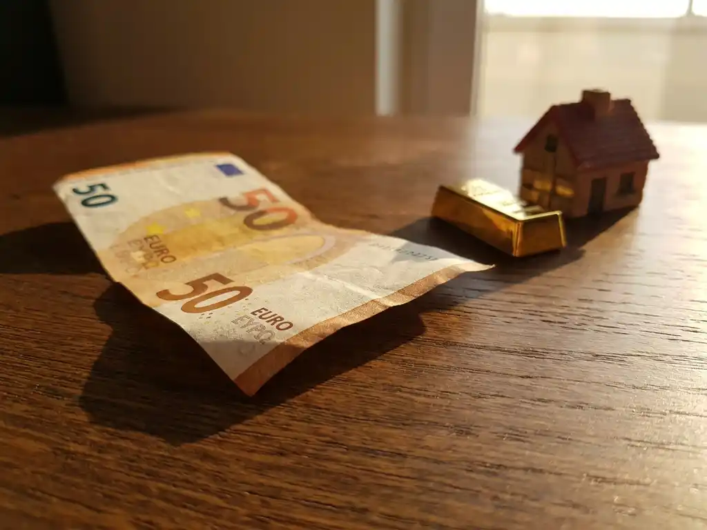 Euro-Geldschein mit sich kräuselnden, gebräunten Rändern auf Eichentisch, daneben Goldbarren und Hausmodell als Inflationsschutz.